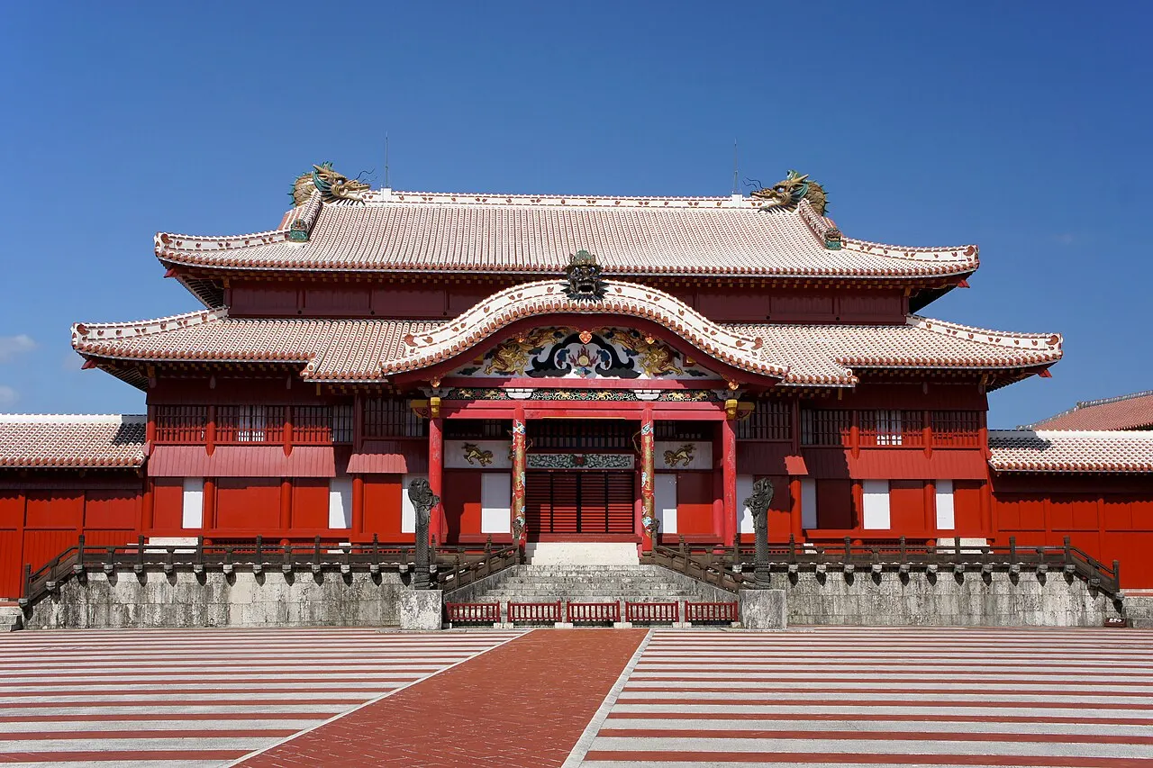The red facade of the Seiden, the main hall of Shuri Castle, with a gabled roof and curved eaves, facing a courtyard of red and white stripes under a blue sky.