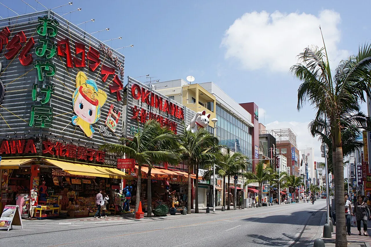 Kokusai-dori on a sunny day, with souvenir shops under colorful signs, palm trees on the sidewalk and buildings along the street.