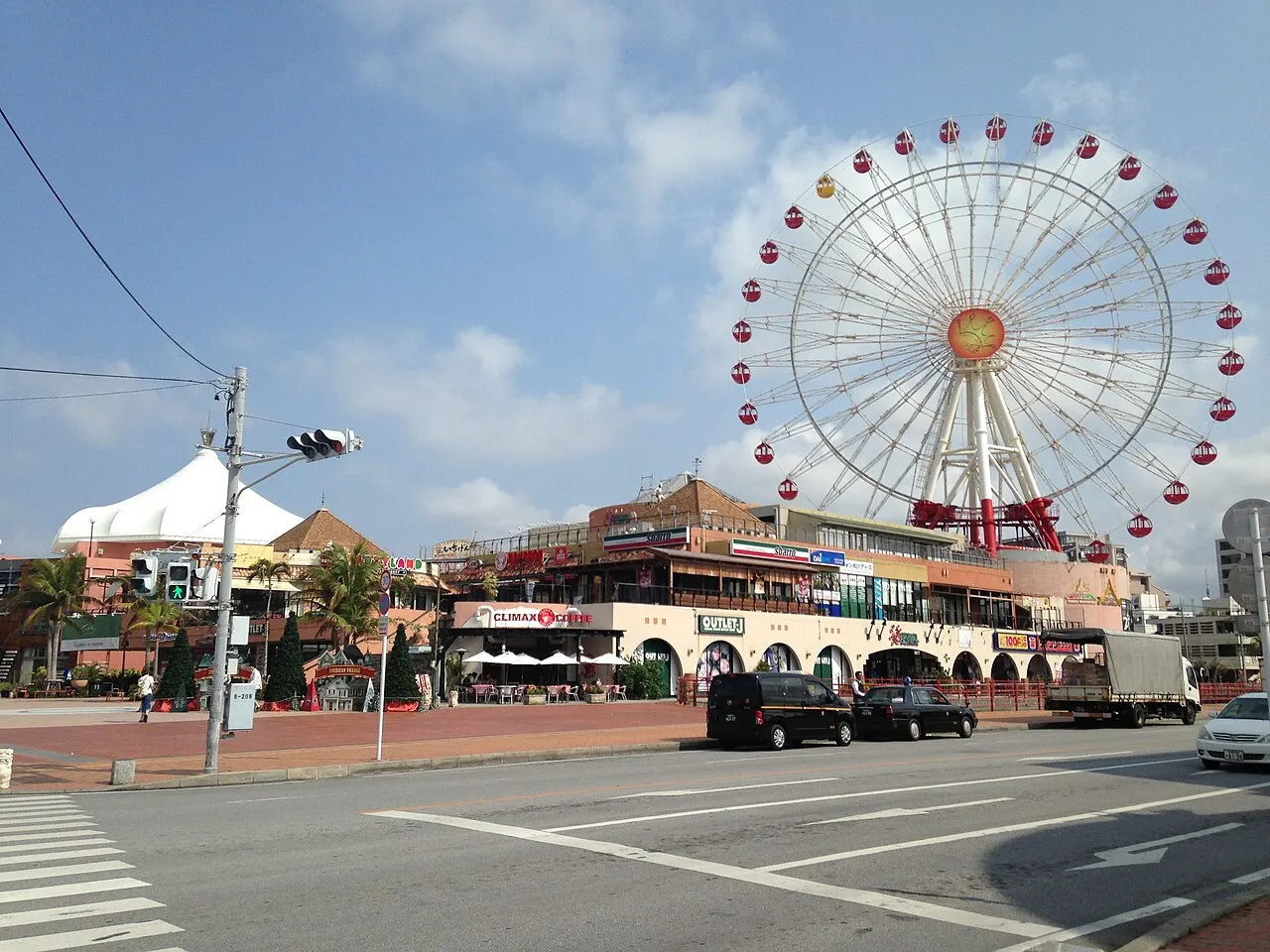 The red and white Ferris wheel of Mihama American Village behind colorful shop buildings, seen from across an intersection.