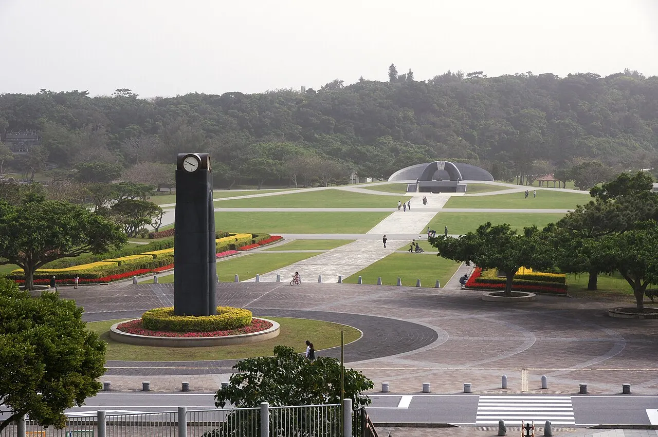 Wide view of the Peace Memorial Park, with a dark clock tower in a round plaza, flower beds, lawns and a domed memorial in the distance.