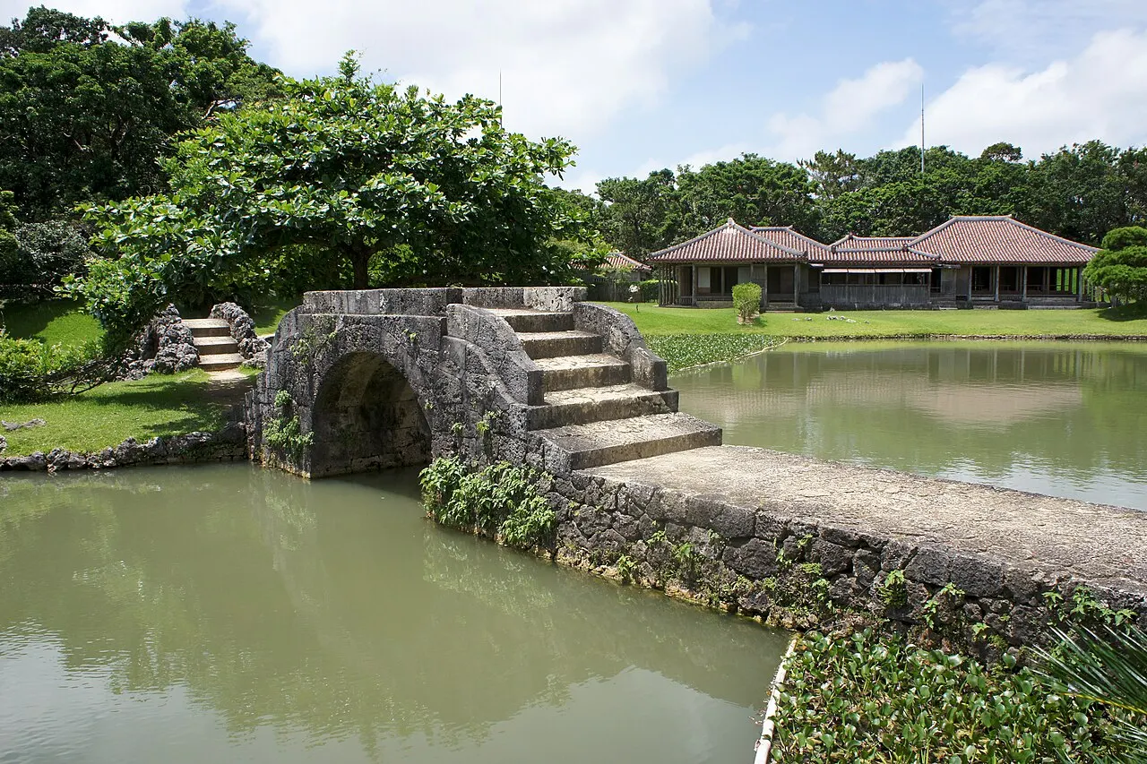 An arched stone bridge over the pond of Shikinaen garden, with the red-roofed pavilion behind among trees.