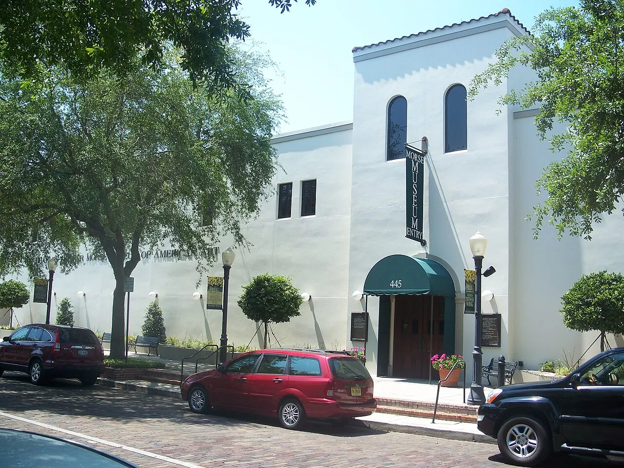 A fachada branca do Charles Hosmer Morse Museum of American Art, em Winter Park, com toldo verde na entrada, árvores e carros estacionados na rua.