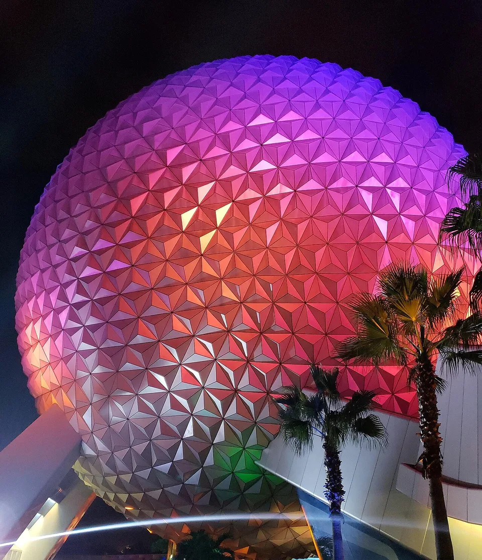 Vista de baixo da esfera Spaceship Earth no EPCOT em novembro de 2018, iluminada em rosa, roxo e laranja, com palmeiras à frente e parte da borda direita fora do quadro.