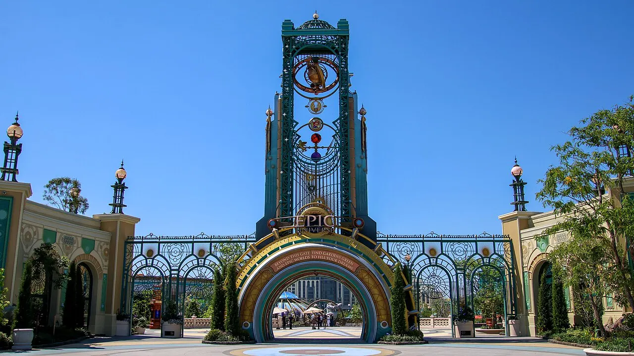 O portal de entrada do Epic Universe, com um arco dourado, grades decoradas e a torre Chronos, alta e esverdeada, sob céu azul.