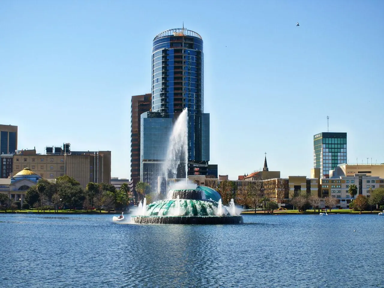 A fonte no meio do Lake Eola jorrando água, com os prédios altos do centro de Orlando atrás e o lago azul em primeiro plano.