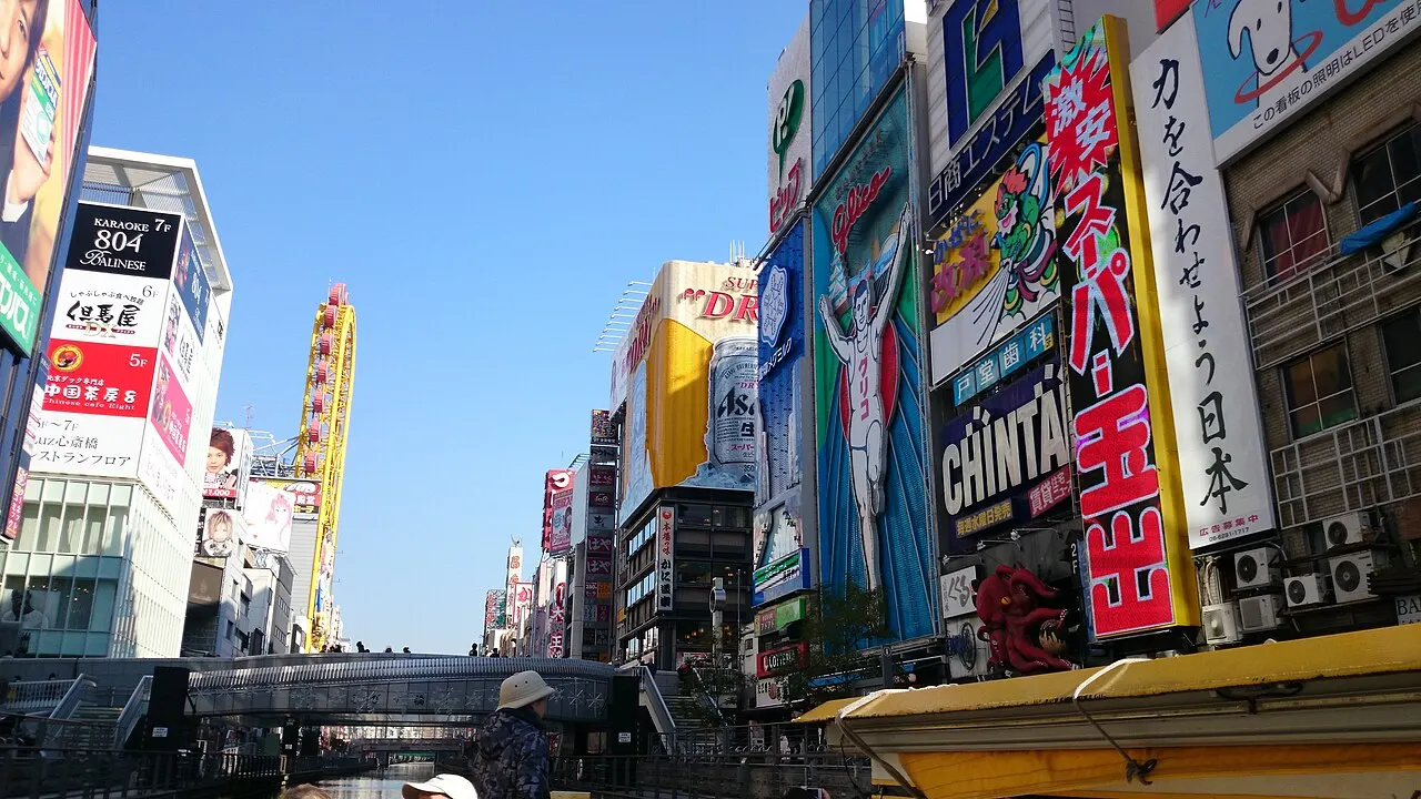 Canal do Dōtonbori em Osaka entre prédios cobertos de letreiros gigantes, entre eles o corredor da Glico e um polvo de propaganda, com uma ponte de pedestres ao fundo.