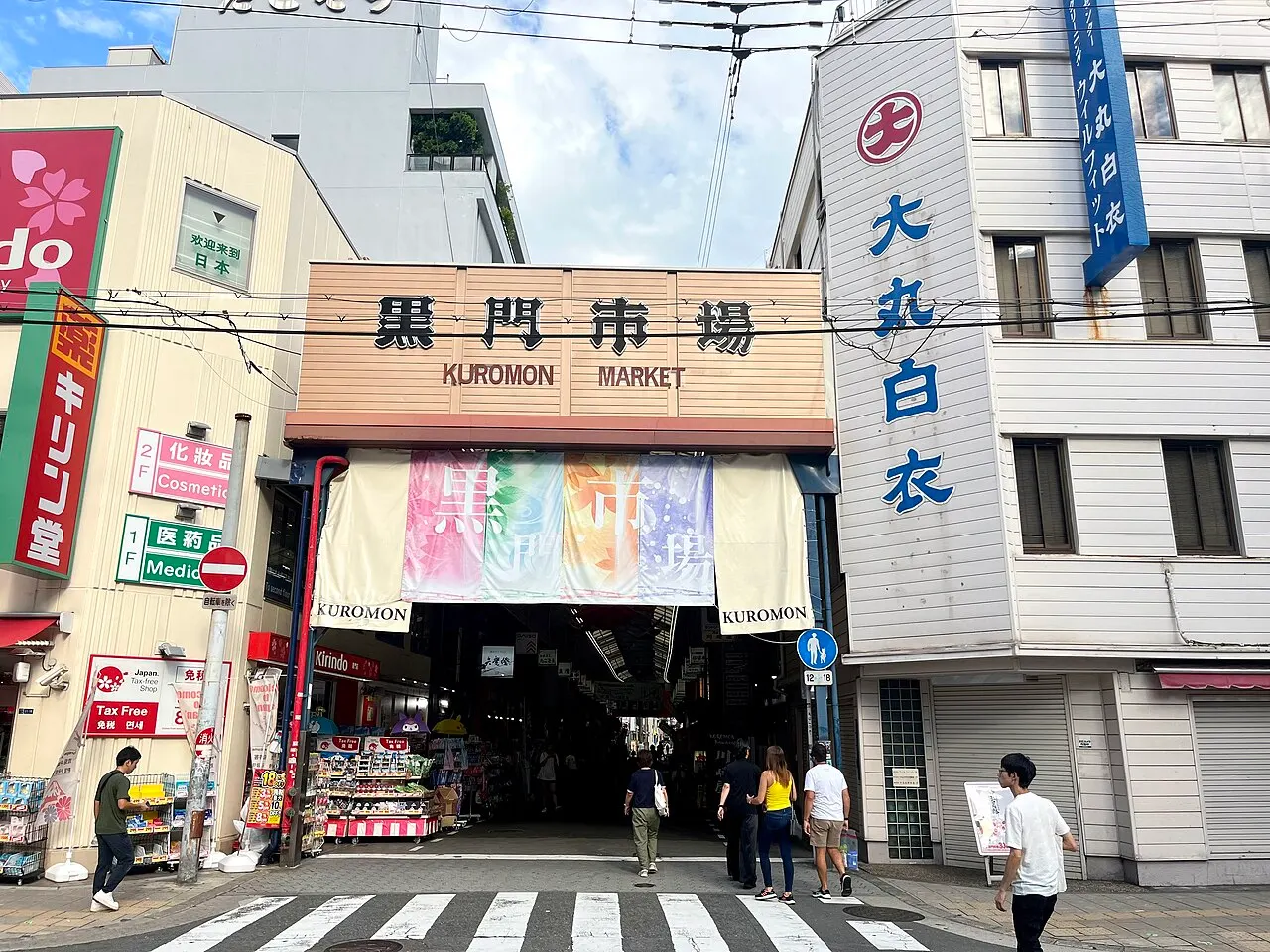 Entrada coberta do Mercado Kuromon, com letreiro de madeira em japonês e inglês, bandeirolas coloridas penduradas e pessoas atravessando a faixa de pedestres.