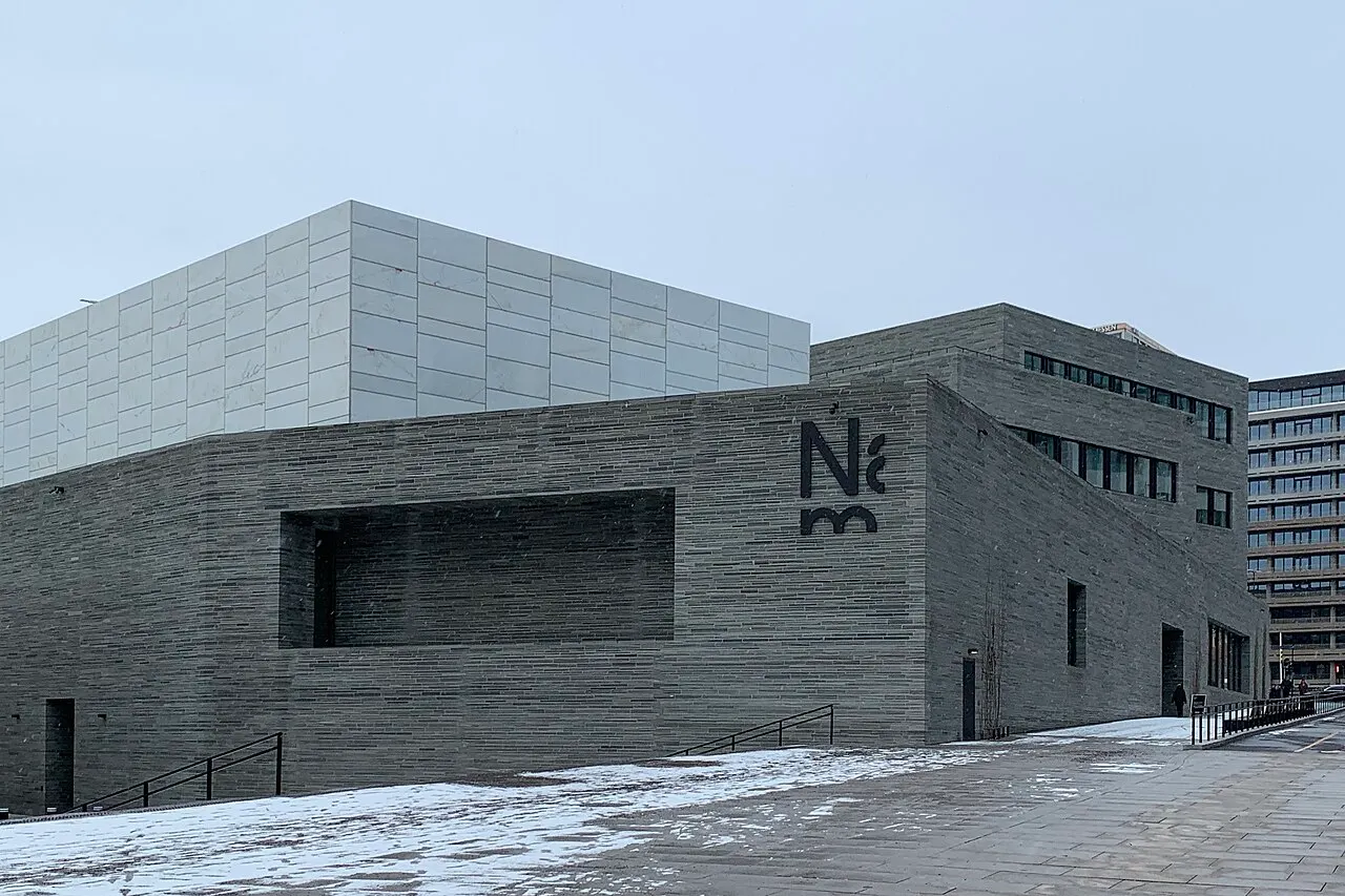Prédio do Nasjonalmuseet, de pedra cinza, com o volume superior de placas claras, visto da rua com neve.