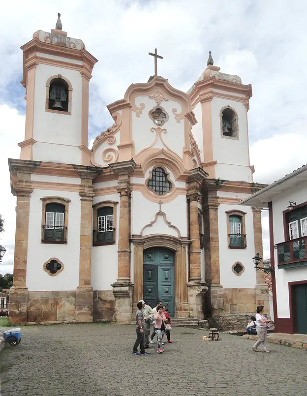 A fachada da Basílica de Nossa Senhora do Pilar, em Ouro Preto, branca com detalhes de pedra, duas torres e pessoas no adro.