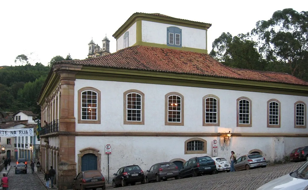 A Casa dos Contos, em Ouro Preto, um sobrado branco de esquina com janelas em arco, telhado de telha e carros estacionados na rua inclinada.