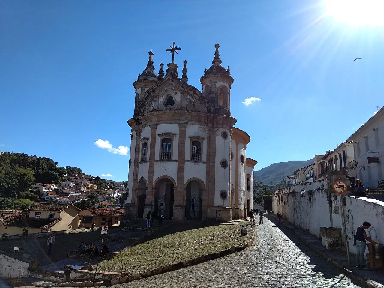 A Igreja de Nossa Senhora do Rosário dos Pretos, em Ouro Preto, com fachada curva branca e de pedra, duas torres e uma rua de pedra descendo ao lado.