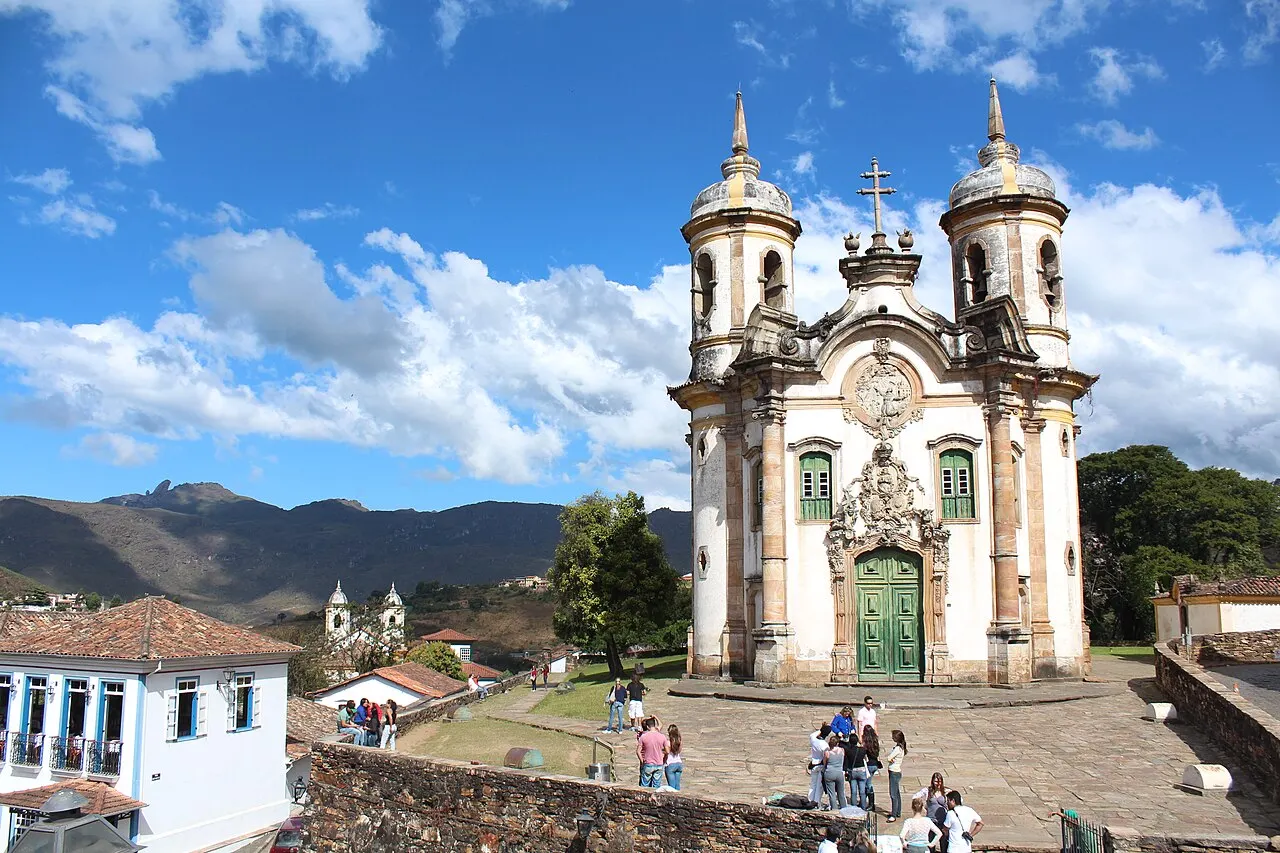 A Igreja de São Francisco de Assis, em Ouro Preto, com fachada branca e pedra, duas torres redondas e o largo com visitantes, diante das montanhas sob céu azul.