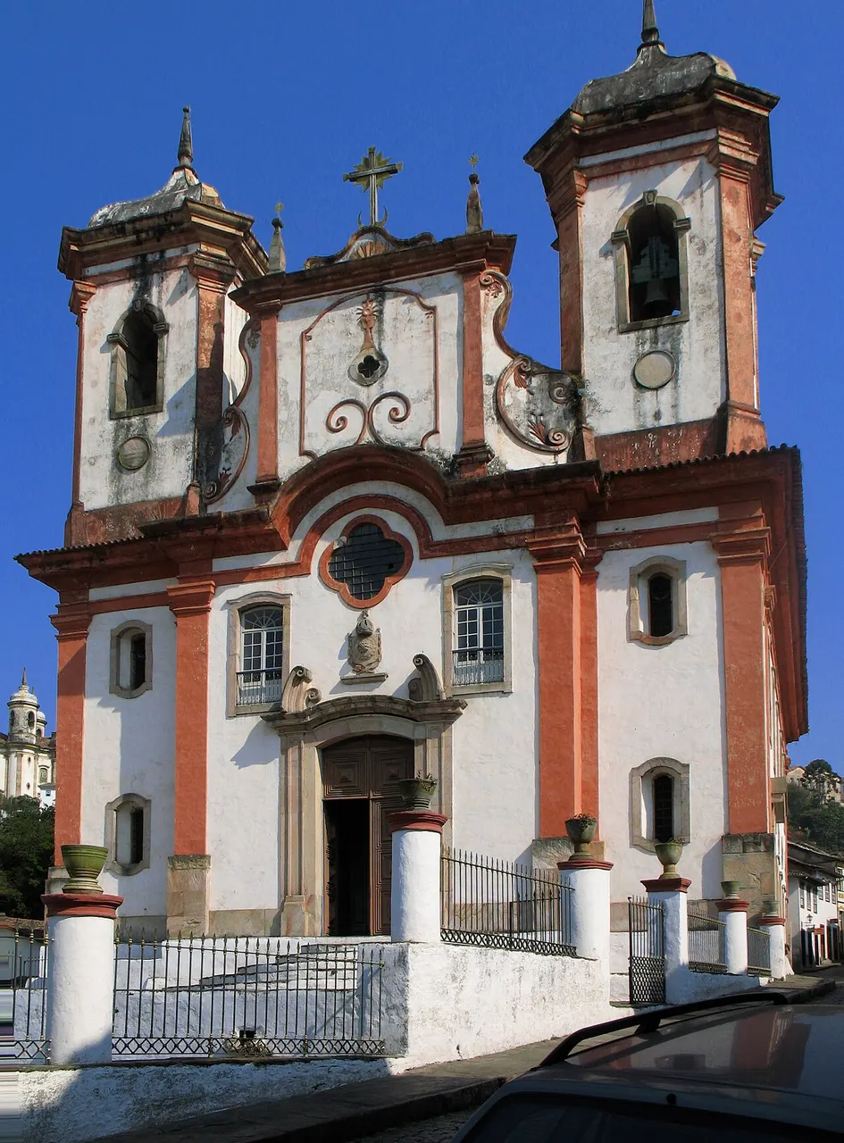 A fachada da Matriz de Nossa Senhora da Conceição de Antônio Dias, em Ouro Preto, branca com contornos avermelhados e duas torres, sob céu azul.