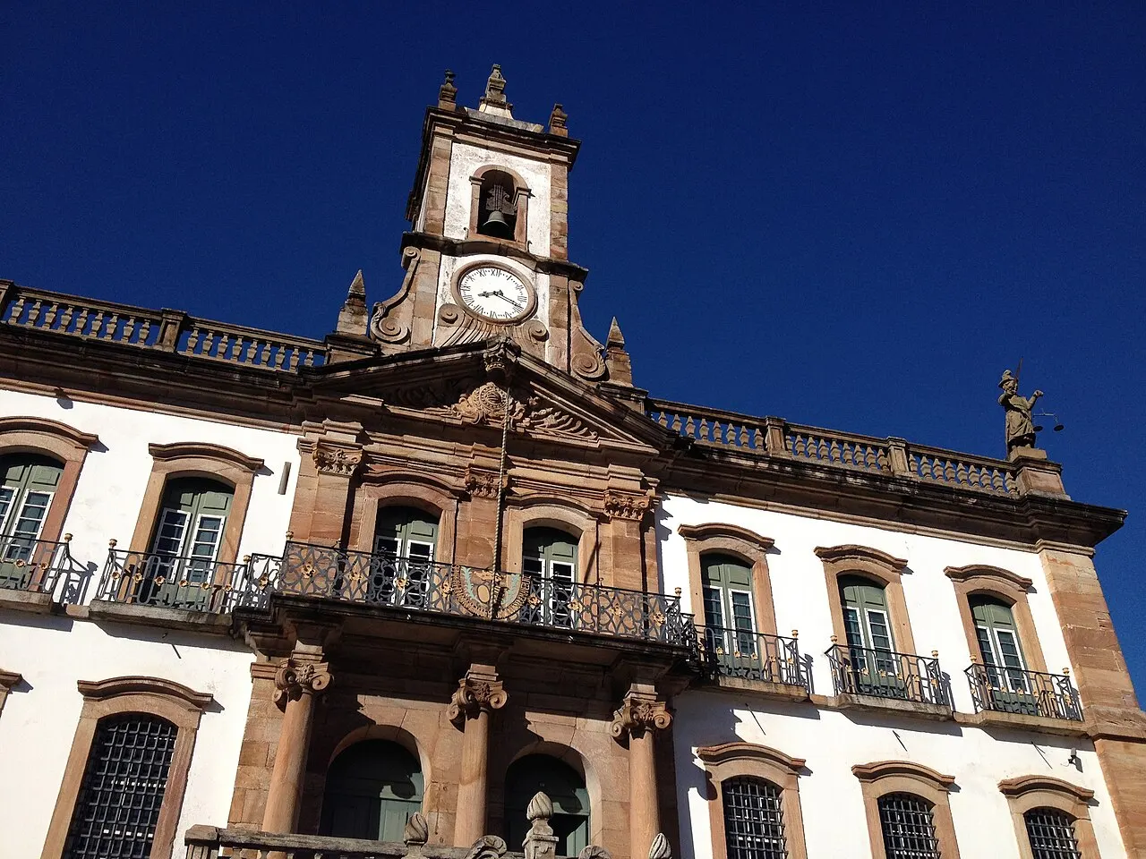 A fachada do Museu da Inconfidência, em Ouro Preto, de pedra e reboco branco, com balcões de ferro e a torre do relógio no alto, vista de baixo contra o céu azul-escuro.