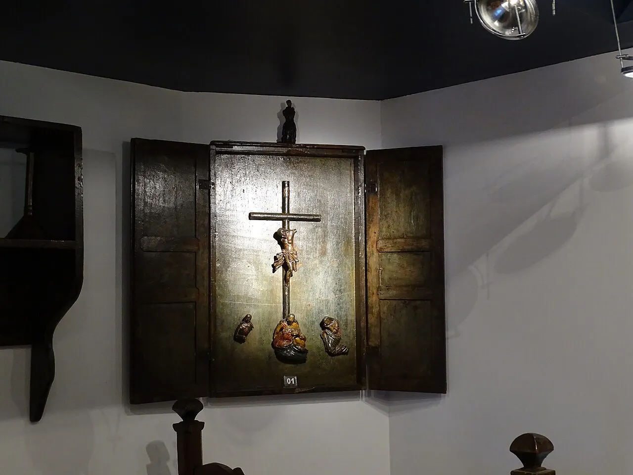 Um oratório de madeira escura com as portas abertas e um crucifixo no interior, exposto na parede do Museu do Oratório, em Ouro Preto.