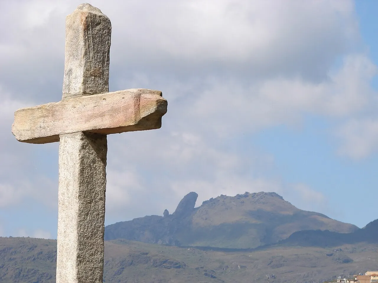 Uma cruz de pedra em primeiro plano e, ao fundo, a serra com o pico do Itacolomi, de formato recortado, sob céu com nuvens.