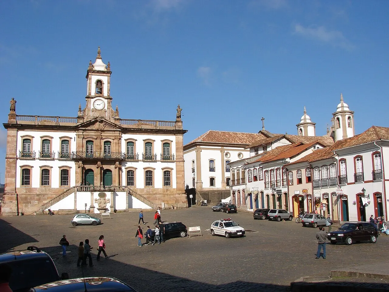 A Praça Tiradentes, em Ouro Preto, com o prédio do Museu da Inconfidência e sua torre do relógio ao fundo, o casario colonial dos lados e carros no calçamento de pedra.