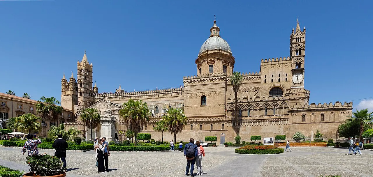 A Catedral de Palermo vista do jardim diante dela, com a cúpula, as torres e as ameias, sob céu azul.