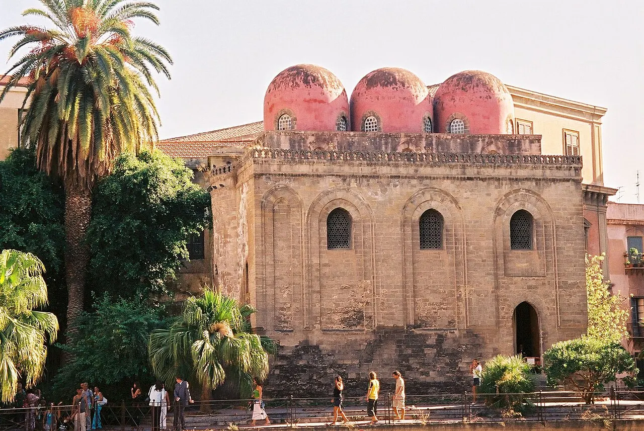 La iglesia de piedra de San Cataldo, cúbica, con tres cúpulas rojas en lo alto y una palmera al lado.