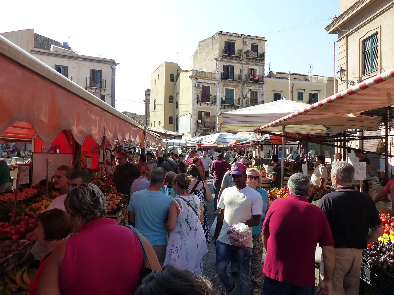 Gente pasando entre los puestos de frutas y verduras del mercado de Ballarò, bajo toldos rojos y edificios antiguos.