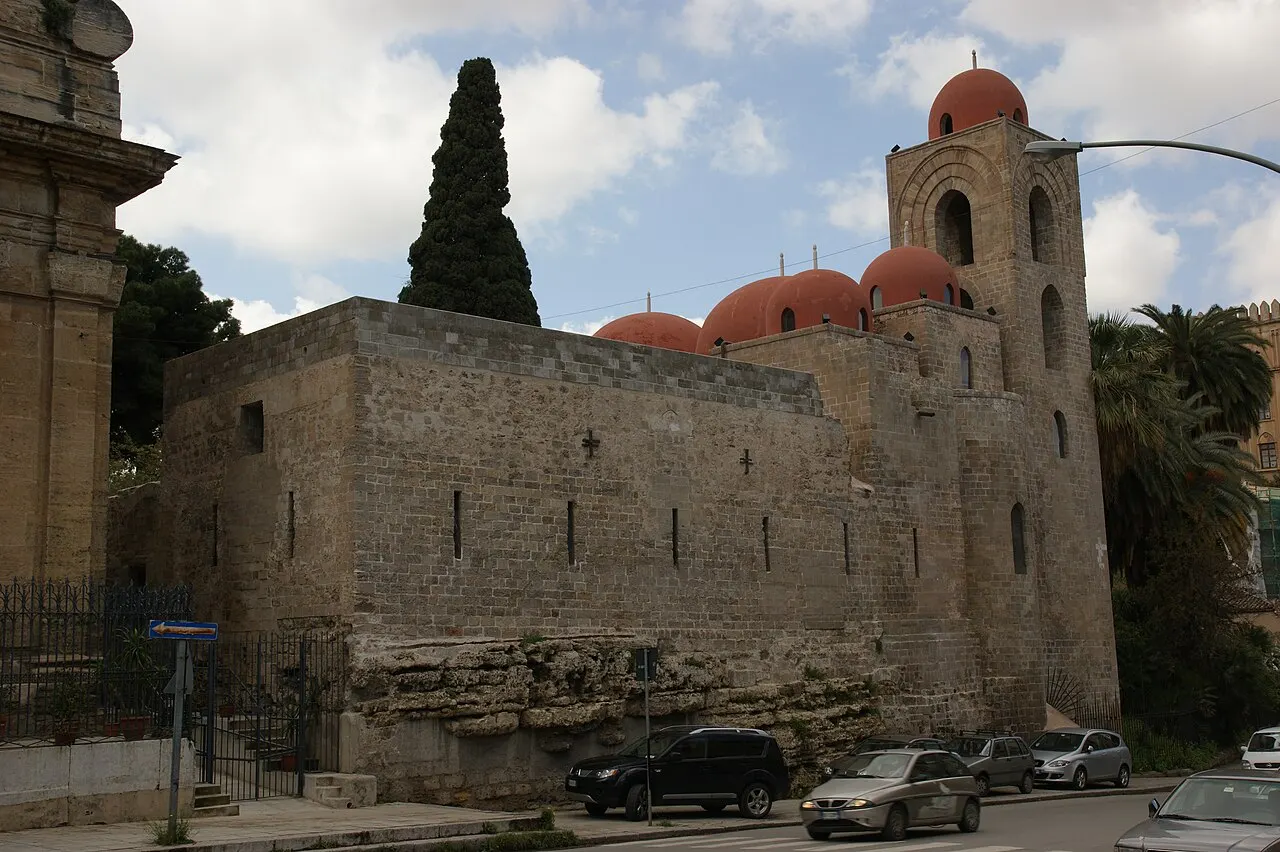 A igreja de San Giovanni degli Eremiti vista da rua: muro de pedra, o campanário e as cúpulas vermelhas entre árvores.