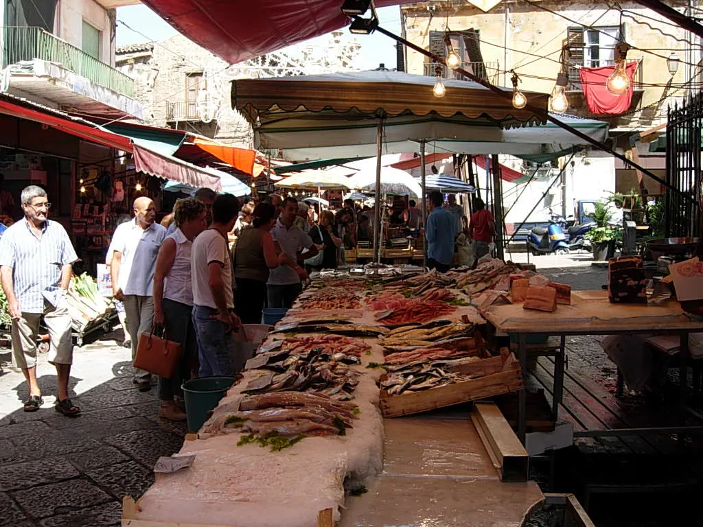 Uma banca de peixe da Vucciria coberta de gelo, sob toldos, com pessoas olhando e passando pela rua estreita.