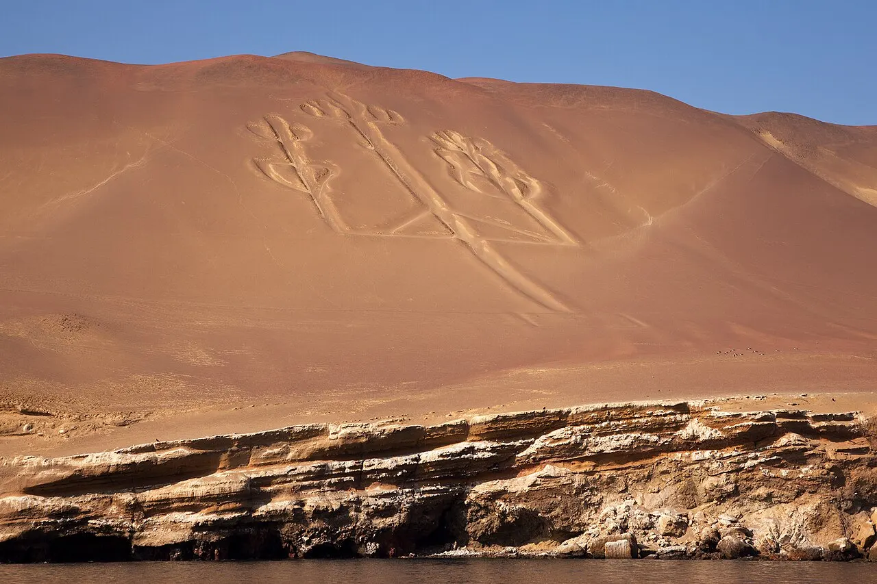 A figura de um candelabro de três braços escavada numa encosta de areia avermelhada, acima de uma faixa de rochas estratificadas à beira da água.