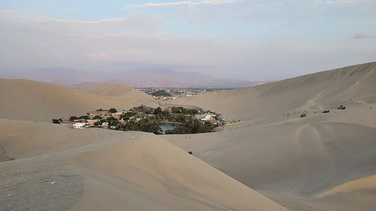 Um pequeno oásis com árvores, construções e uma lagoa verde no fundo de um vale de dunas de areia, com pessoas pequenas no alto das dunas.
