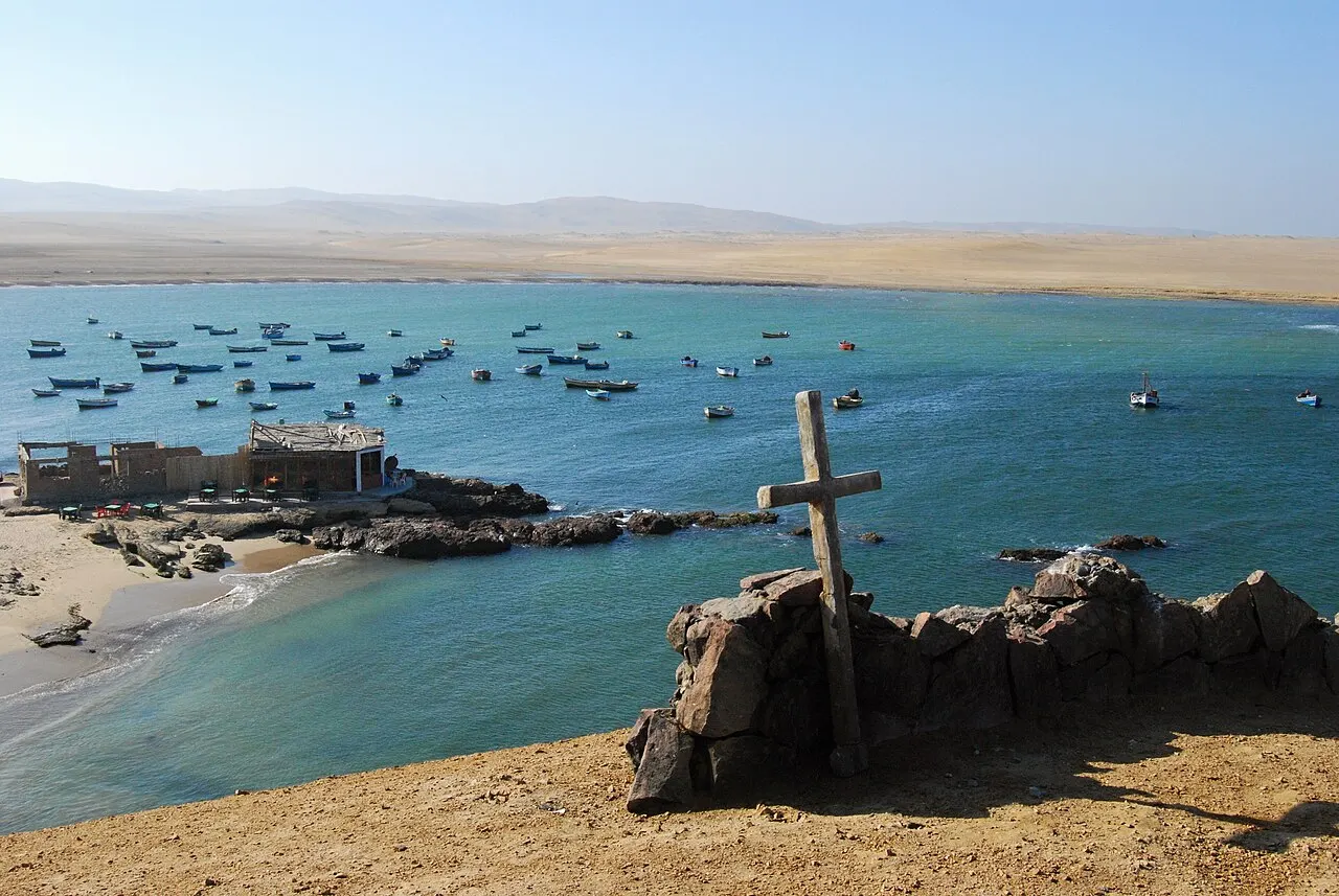 Dezenas de barcos de pesca ancorados numa baía azul, com construções baixas na beira e uma cruz de madeira sobre pedras em primeiro plano.