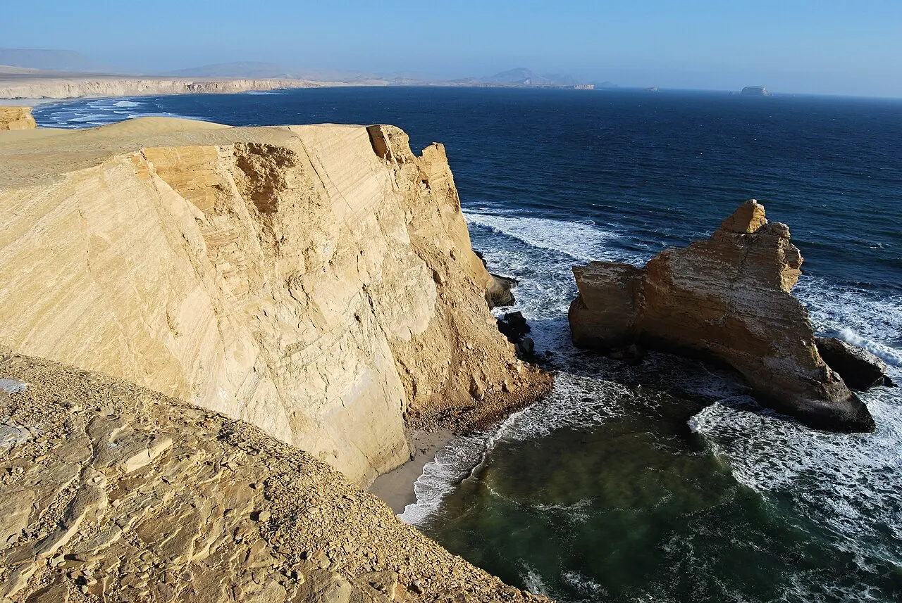 Um rochedo isolado de pedra clara no mar, com ondas batendo em volta, ao lado de uma falésia alta vista de cima.