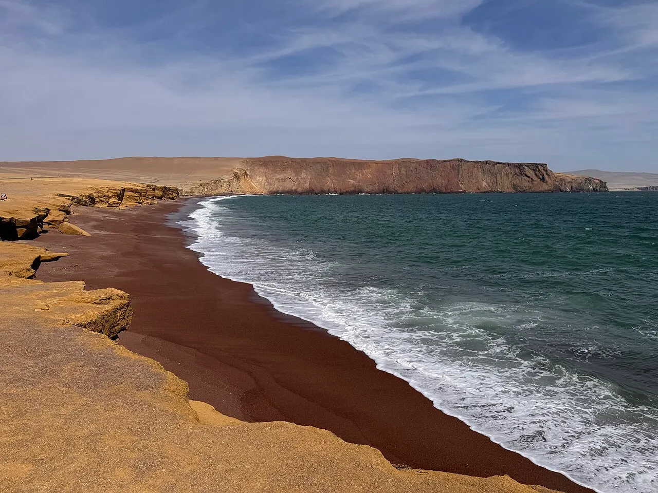 Uma praia de areia vermelho-escura em curva, entre o mar verde com ondas e a falésia amarela, com um promontório ao fundo.