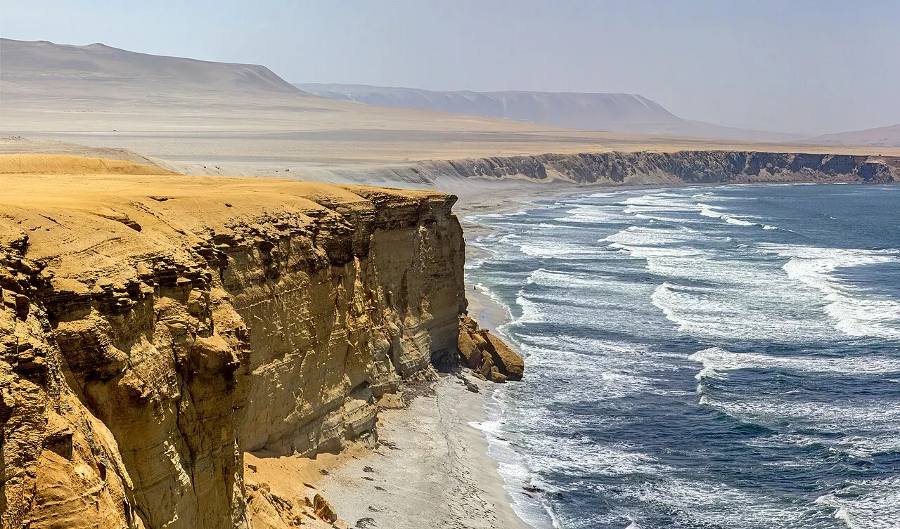 Falésias de areia dourada caindo sobre uma praia estreita, com ondas brancas chegando e montanhas desérticas ao fundo.