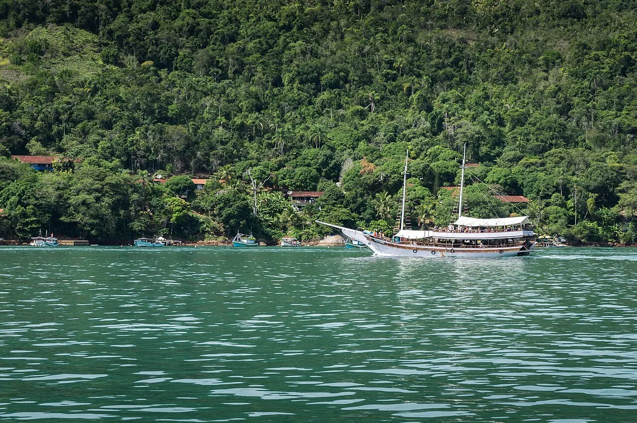 Uma escuna de passeio navegando na água verde da baía de Paraty, diante de uma encosta coberta de mata atlântica.