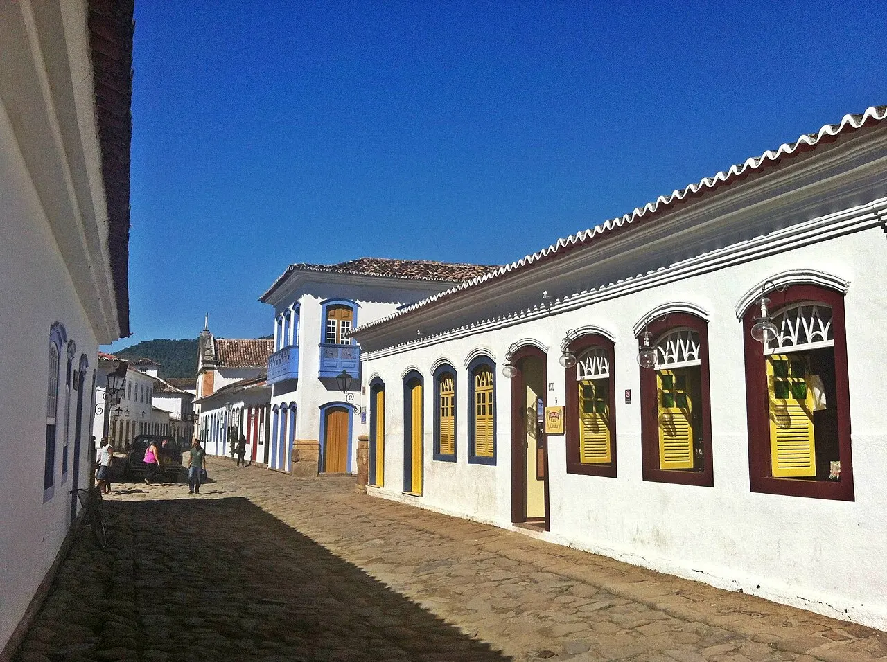 Uma rua de pedras irregulares no centro histórico de Paraty, entre casas coloniais brancas com portas e janelas de molduras coloridas, sob céu azul.