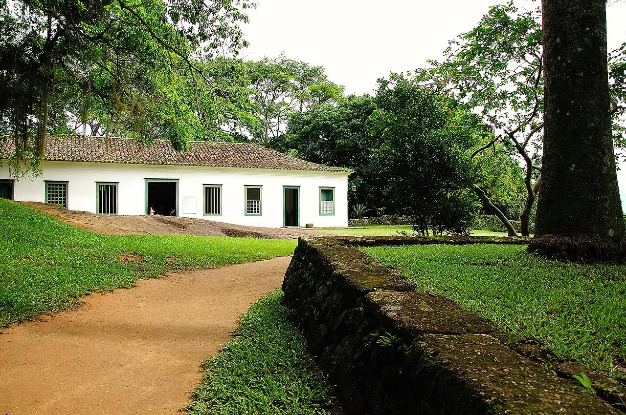 O prédio baixo e branco do Forte Defensor Perpétuo, em Paraty, com janelas de madeira, um caminho de terra, um muro de pedra coberto de musgo e árvores grandes.