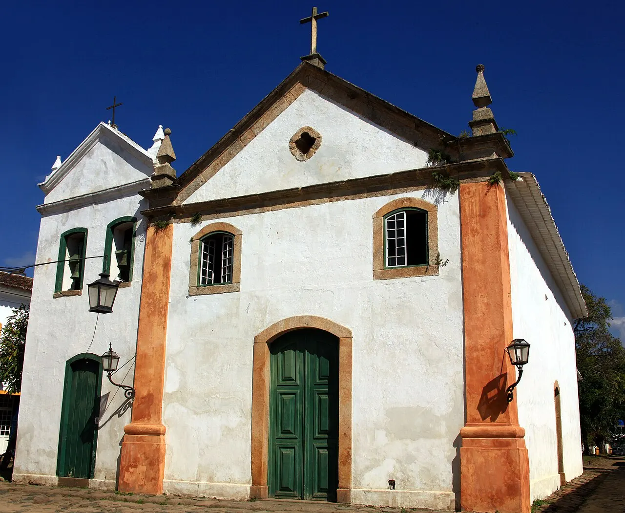 A Igreja de Nossa Senhora do Rosário e São Benedito, em Paraty, branca com cantos alaranjados, porta verde e uma cruz no topo, contra o céu azul.