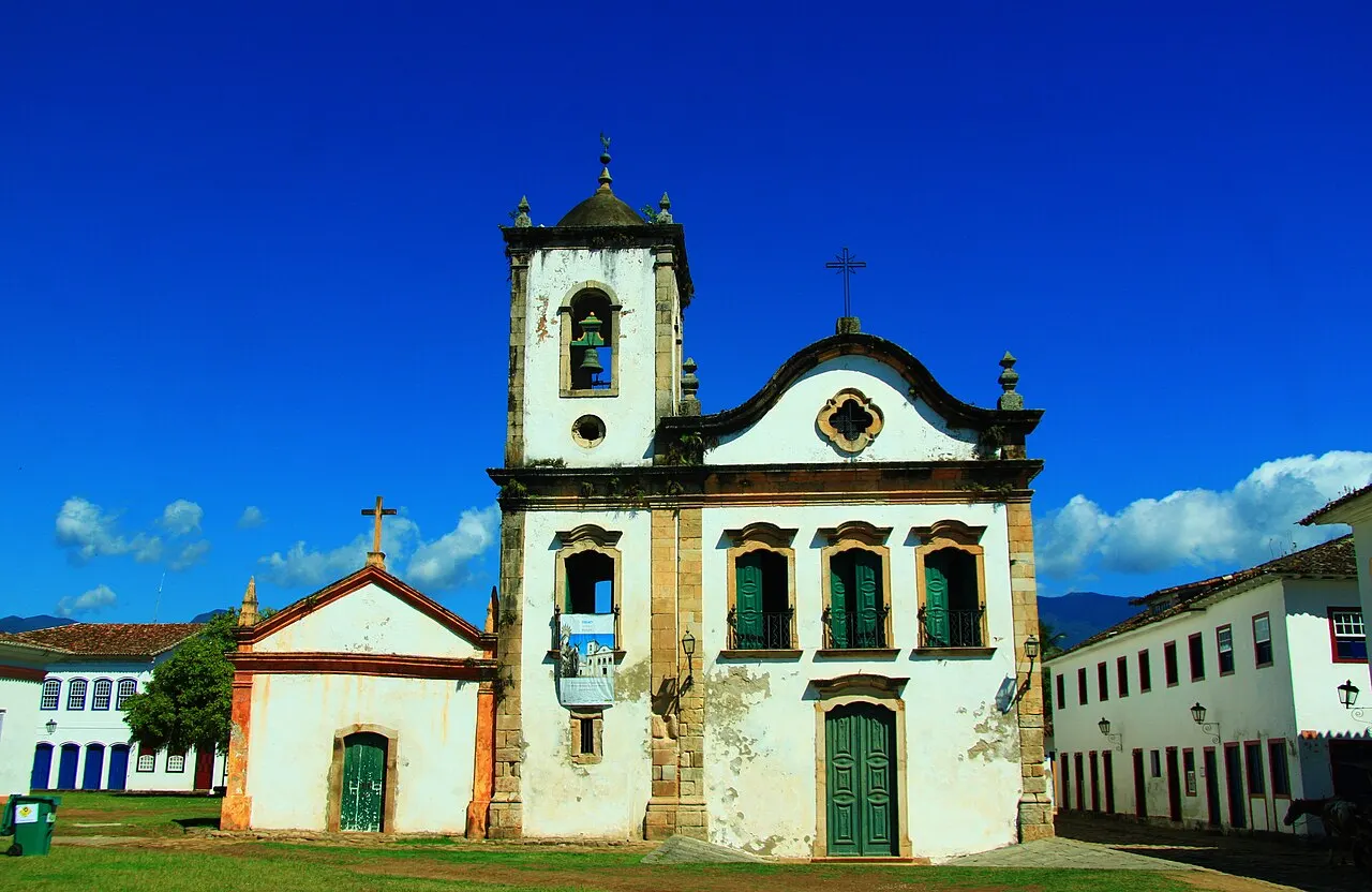 A Igreja de Santa Rita, em Paraty, branca com contornos amarelos, uma torre sineira e uma pequena capela ao lado, sobre o gramado, sob céu azul intenso.