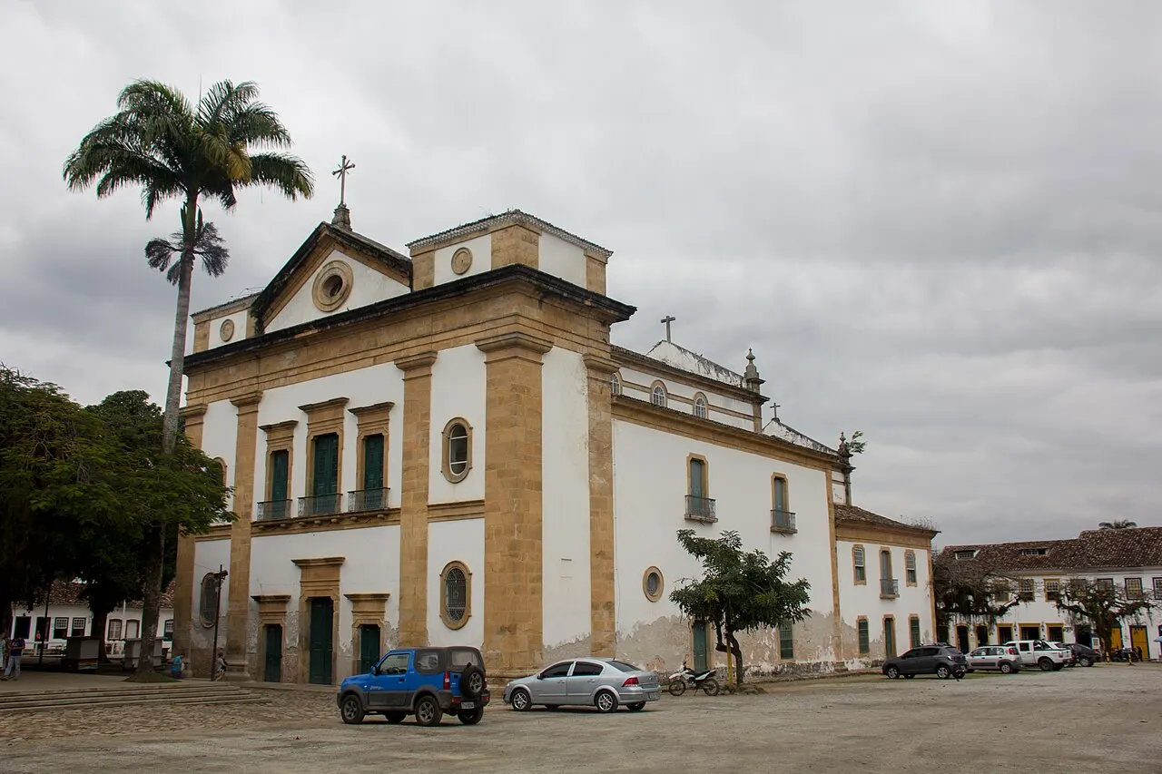 A Igreja Matriz de Nossa Senhora dos Remédios, em Paraty, de fachada clara com pilastras de pedra, ao lado de palmeiras e carros estacionados na praça, sob céu nublado.
