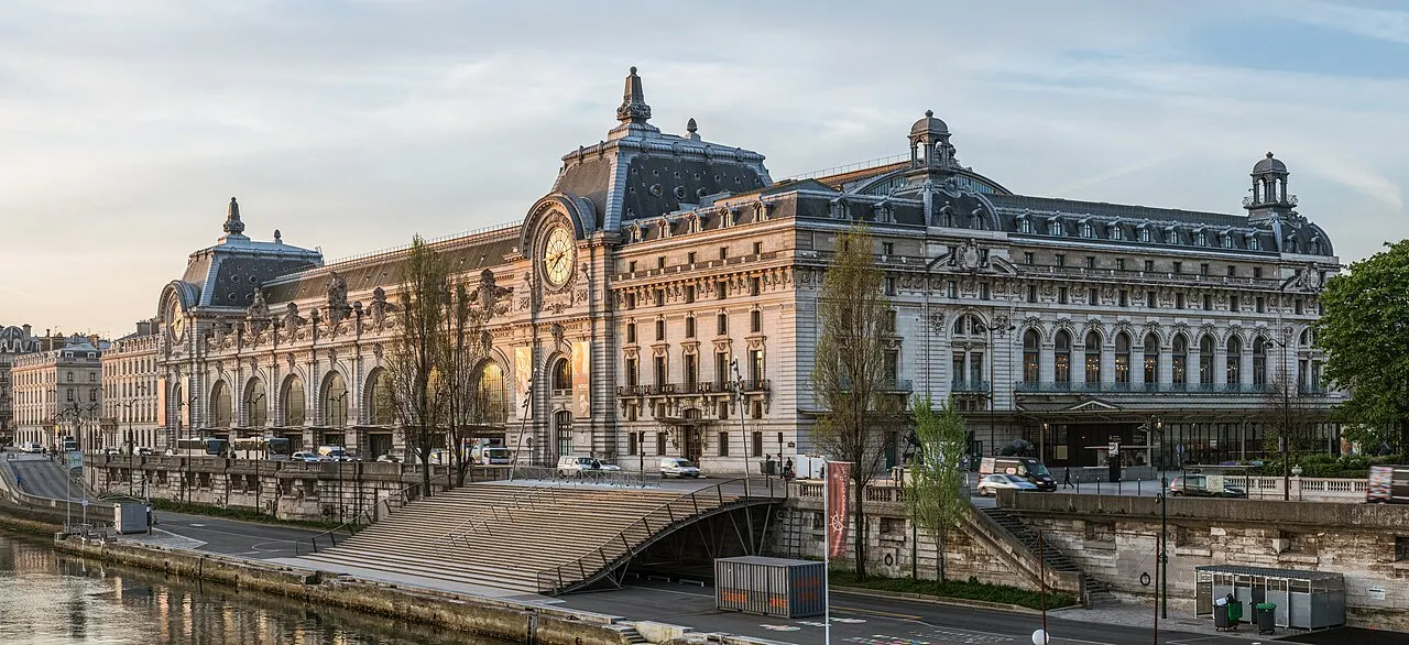A antiga estação de Orsay, longa fachada de pedra com relógios e cúpulas, em luz de fim de tarde à beira do Sena.