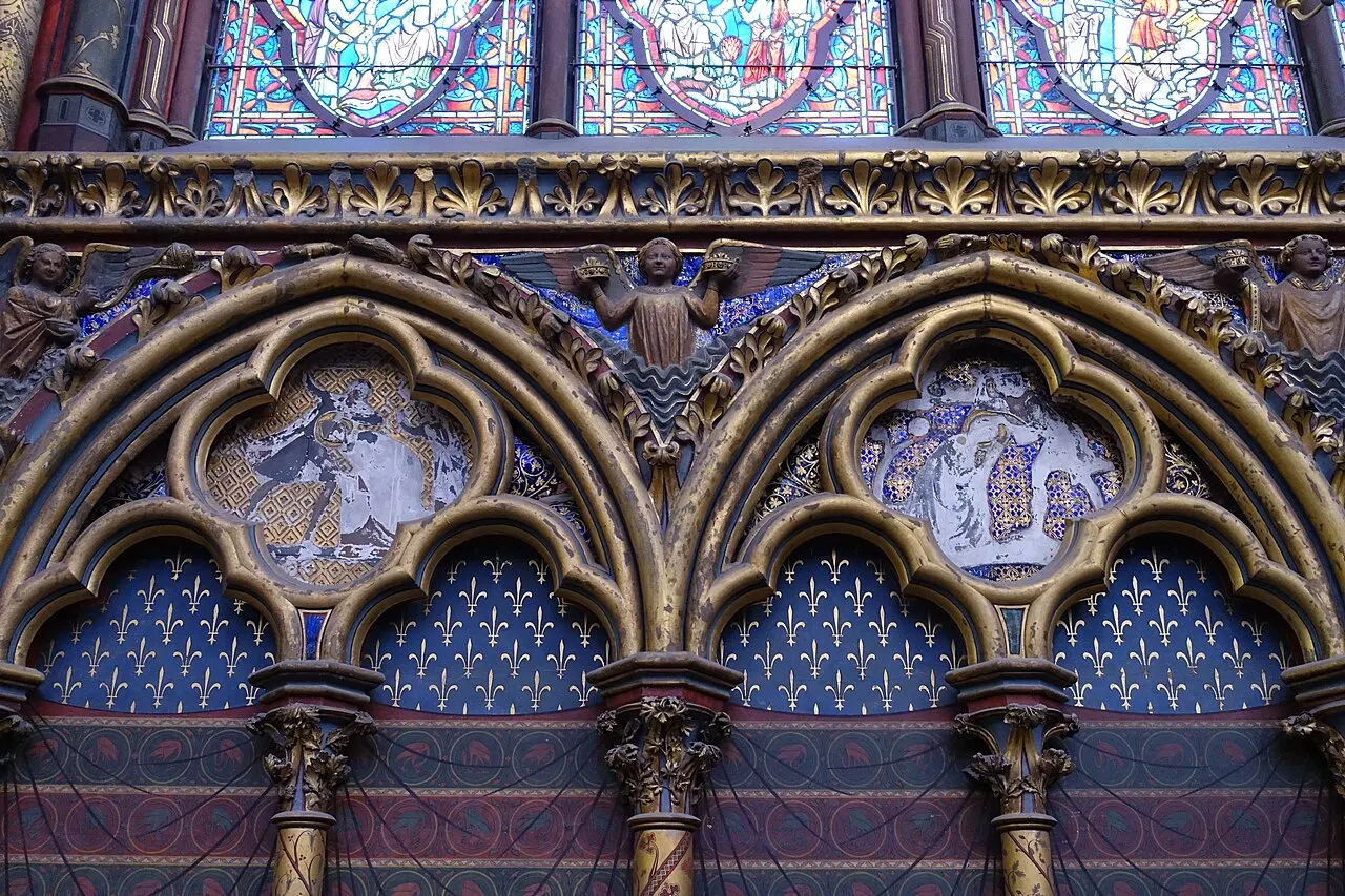 Arcadas góticas pintadas de azul con flores de lis doradas en la capilla alta de la Sainte-Chapelle, medallones esculpidos, un ángel coronado en el centro y el pie de las vidrieras arriba.
