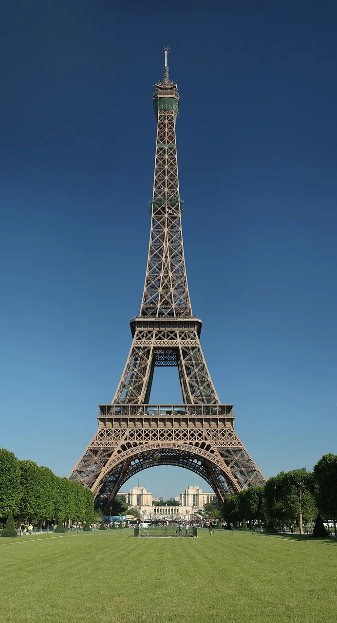Torre Eiffel inteira contra o céu azul, com o gramado do Champ-de-Mars à frente.