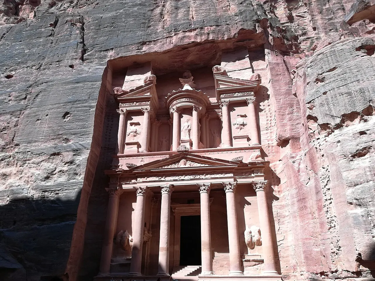 Fachada rosada de Al-Khazneh talhada na rocha do desfiladeiro, com colunas coríntias, frontão triangular e uma rotunda esculpida acima da entrada, sob luz de sol direta.