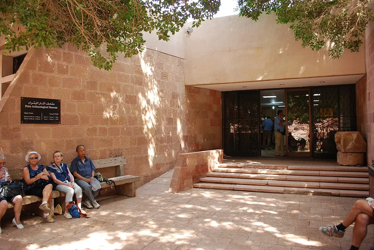 Pátio de pedra com a placa do museu e a porta de vidro, sob uma árvore.
