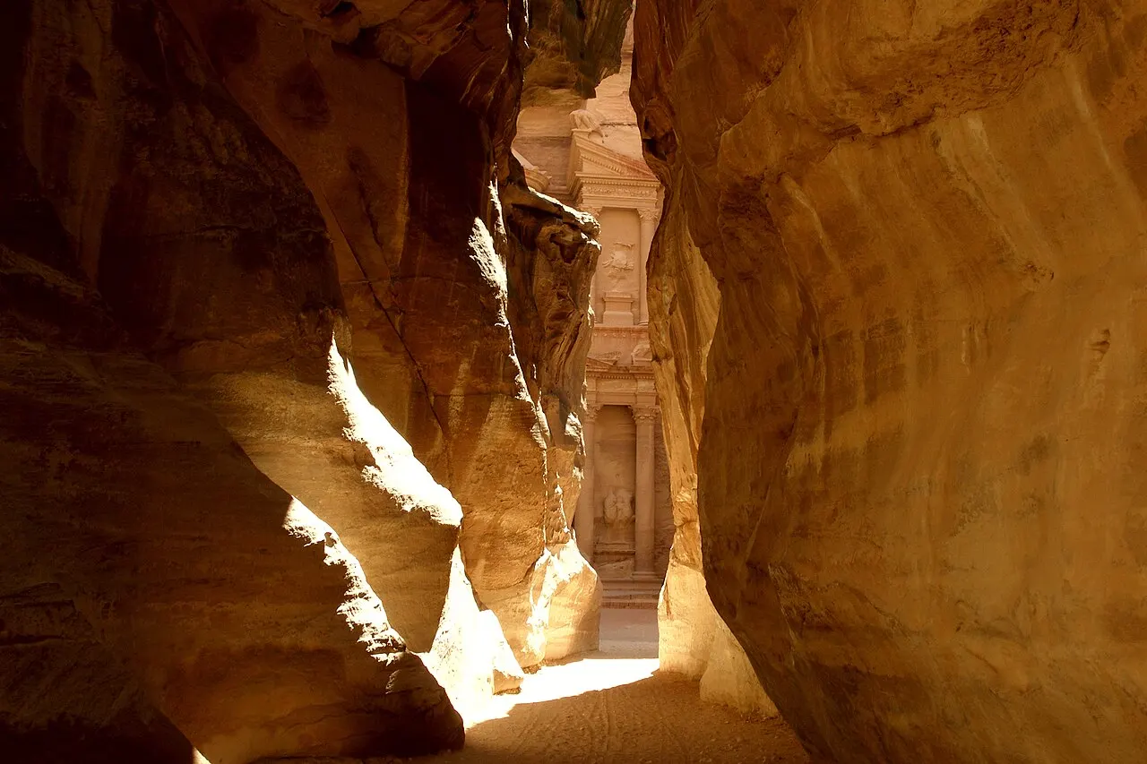 Corredor estreito entre paredões de arenito alaranjado; ao fundo, na fresta, aparece a fachada com colunas do Tesouro de Petra.