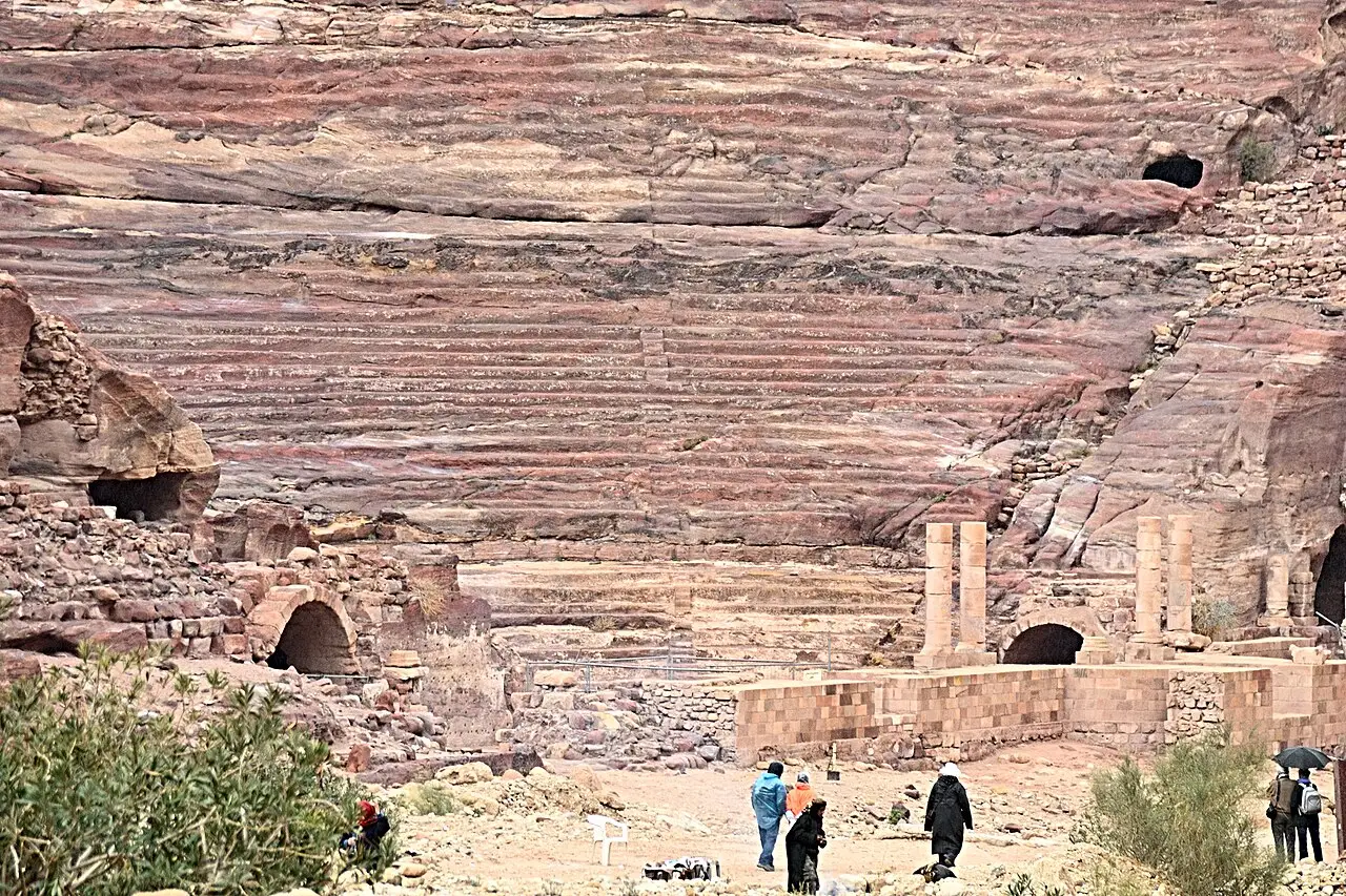 Teatro de Petra talhado na rocha, com a arquibancada, colunas do palco e visitantes na frente.