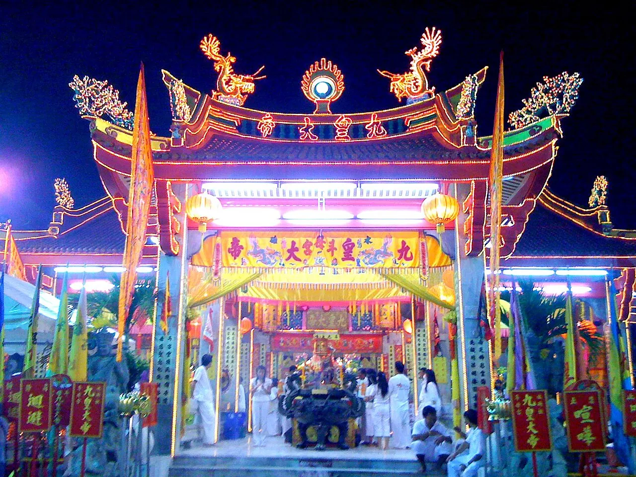 O portal do santuário Jui Tui iluminado à noite, com dragões dourados no telhado, lanternas vermelhas e fiéis de branco.