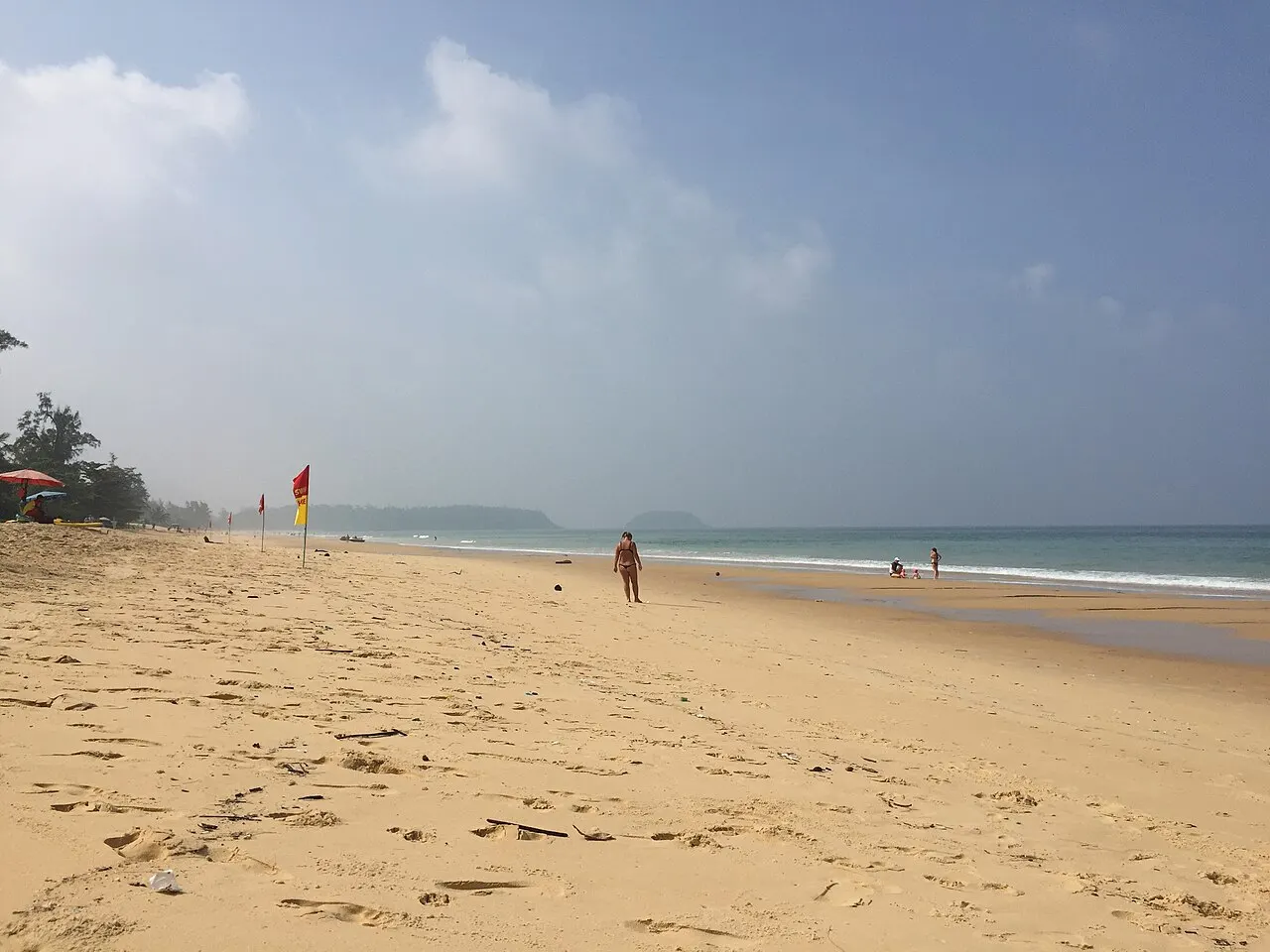 A praia de Karon, uma faixa larga de areia dourada quase vazia, com o mar ao fundo e céu nublado.
