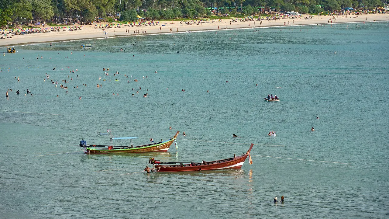 Dois barcos de cauda longa coloridos na água verde e calma de Kata, com banhistas e a faixa de areia ao fundo.
