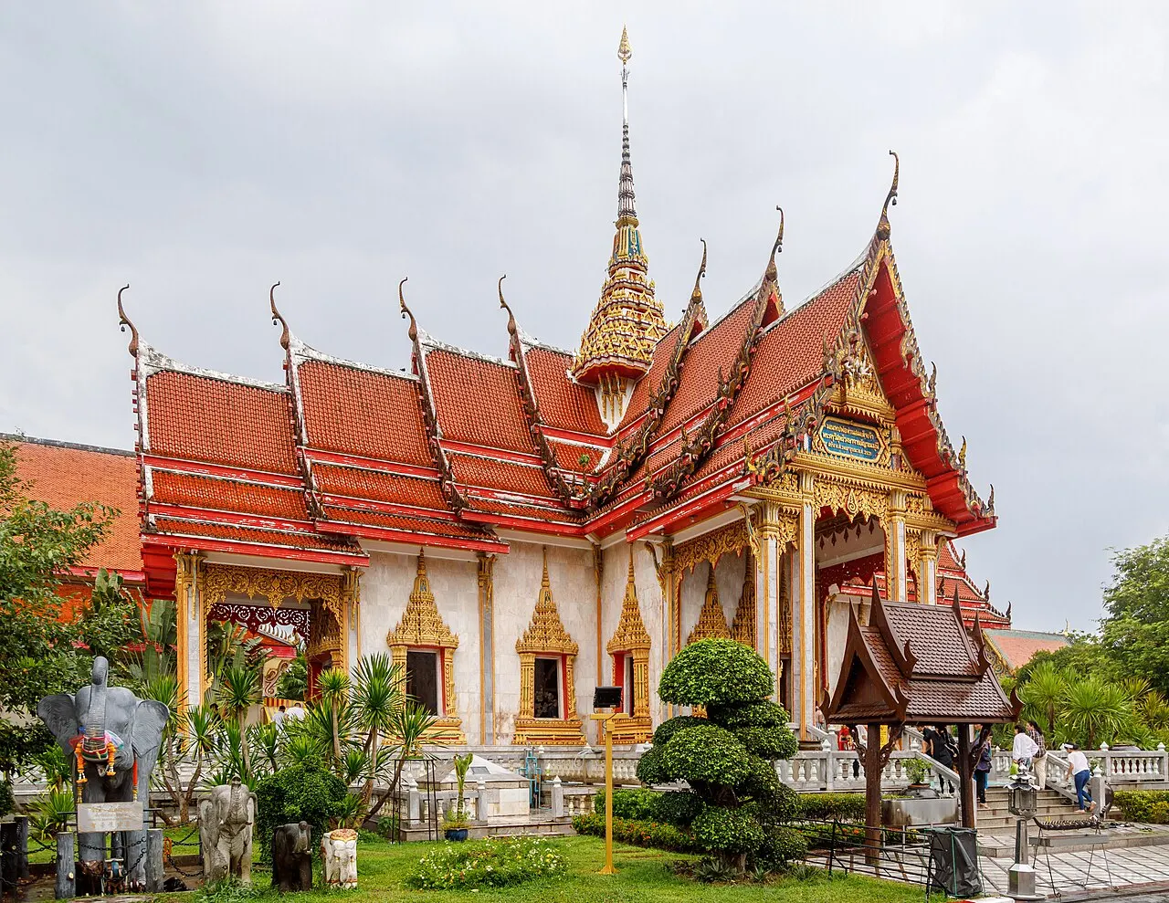 Le pavillon du Wat Chalong, aux toits rouges superposés à bordures dorées et à la flèche pointue, derrière un jardin.