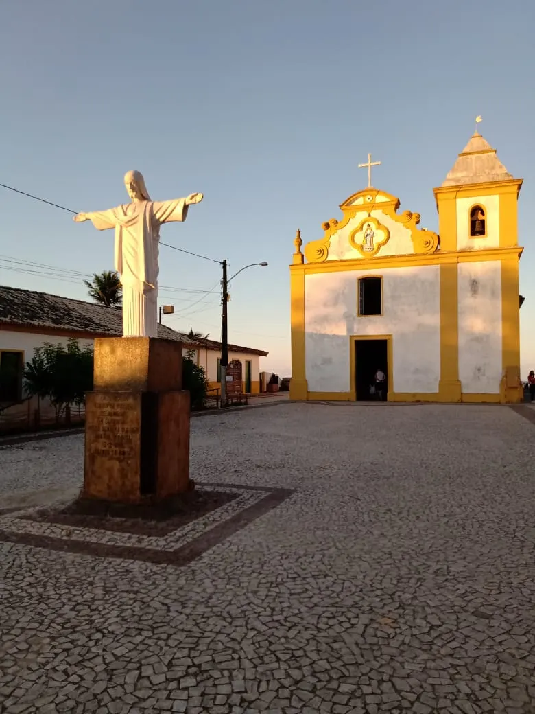 A igreja de Nossa Senhora d'Ajuda, branca com detalhes amarelos e torre, no fim de uma praça calçada, com uma estátua à esquerda ao entardecer.
